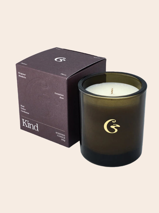Kind scented candle in a dark glass jar next to a purple box listing fragrance notes including bergamot, mandarin, geranium, rose, vetiver, and cardamom.