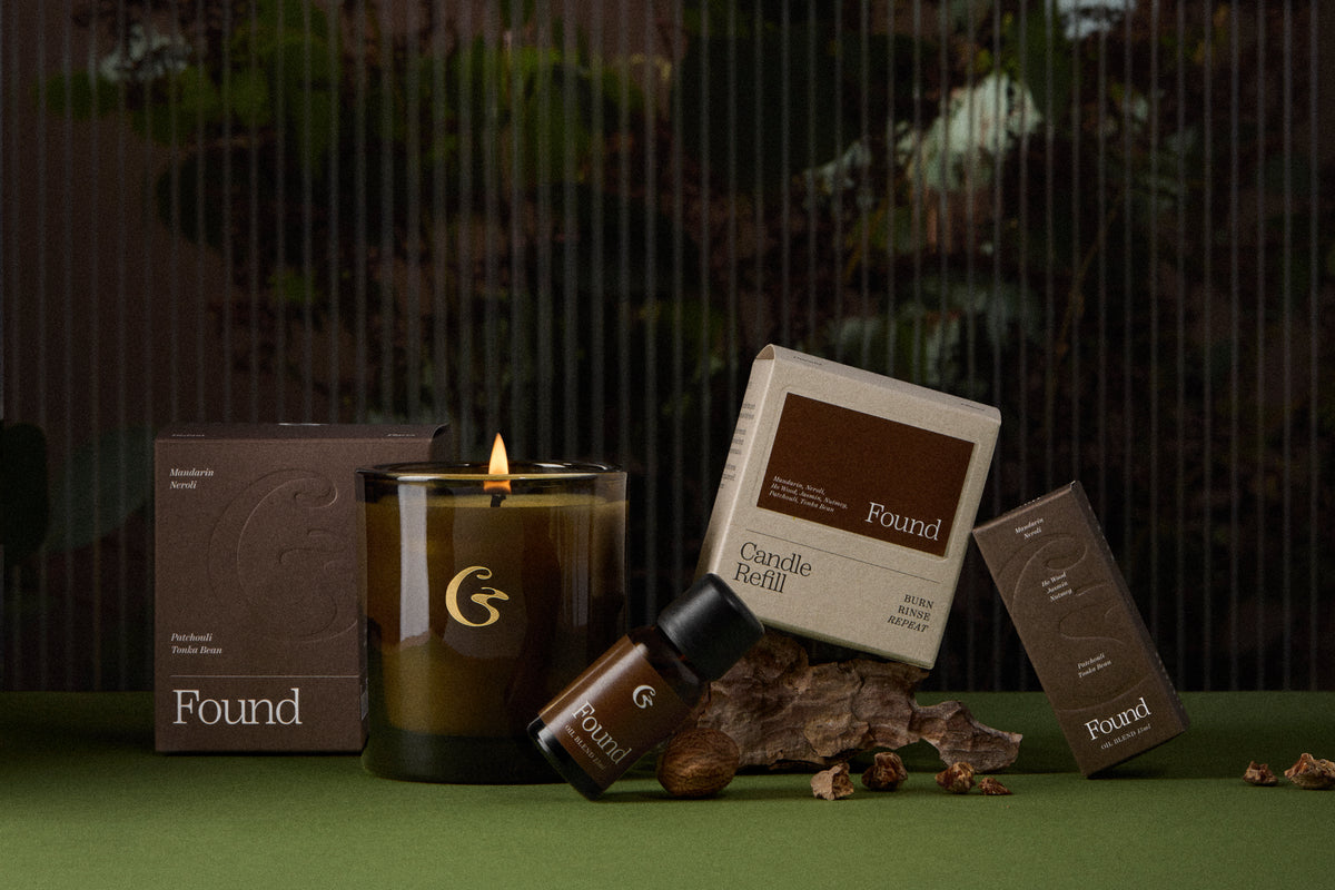 A Found brand candle set arranged on a green surface against a dark, blurred background. The set includes a lit amber glass candle jar with a gold logo, a cream-colored candle refill box, a brown wax melt box, a small brown bottle, and matching brown packaging boxes.