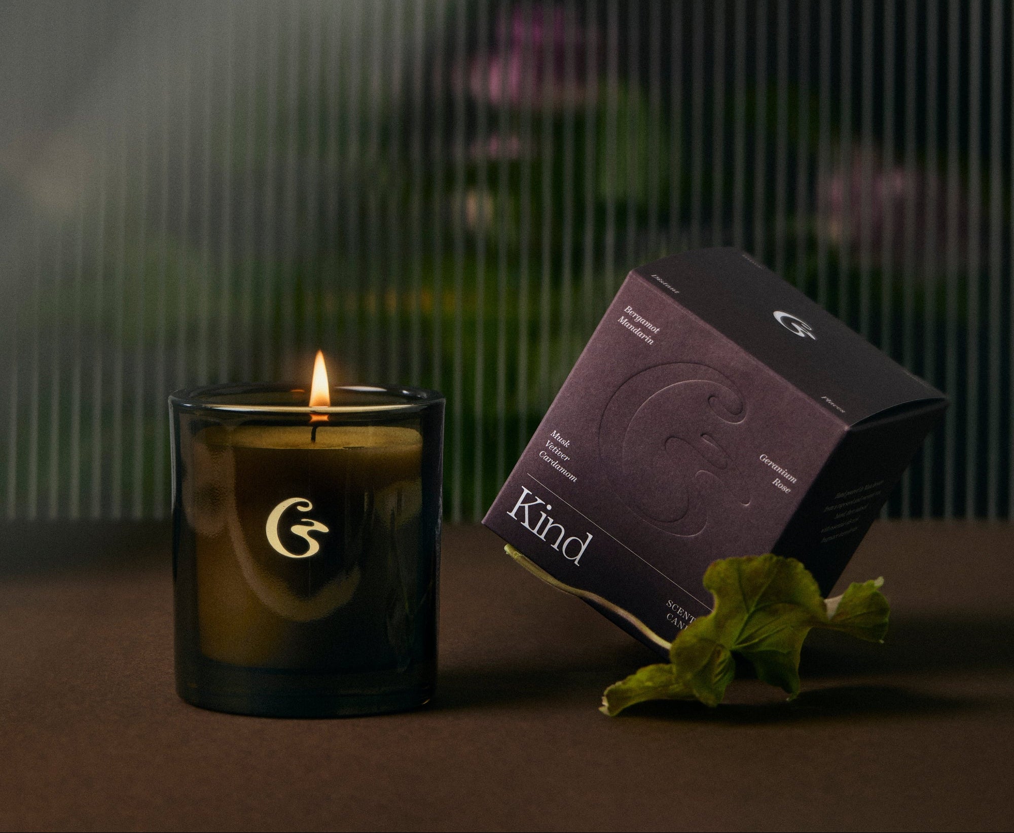 Candle in an amber jar with a purple box with the scent name 'Kind' on there. Floral background