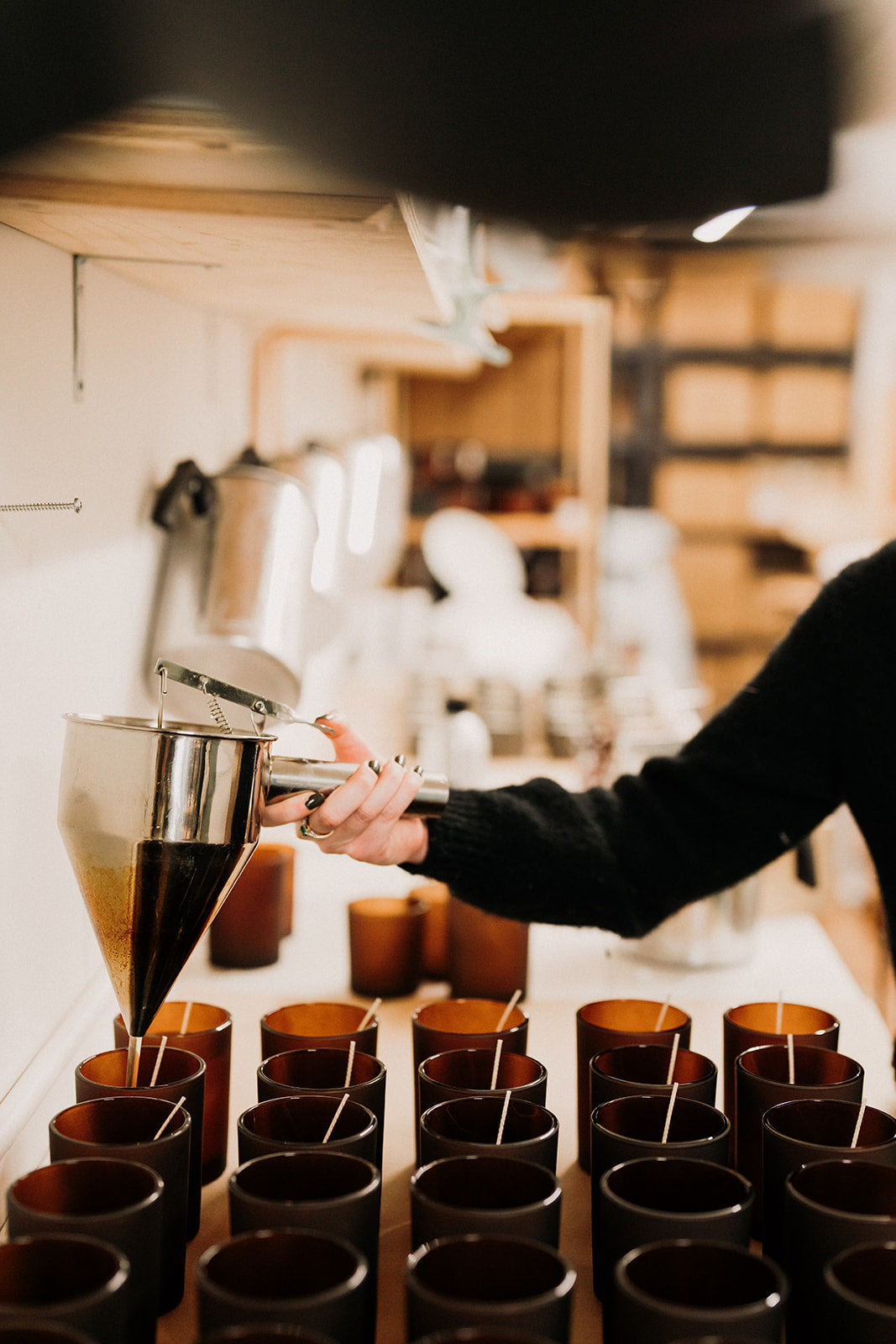 Amber candle jars lined up with wicks in. Wax is being hand poured into the jars in a studio
