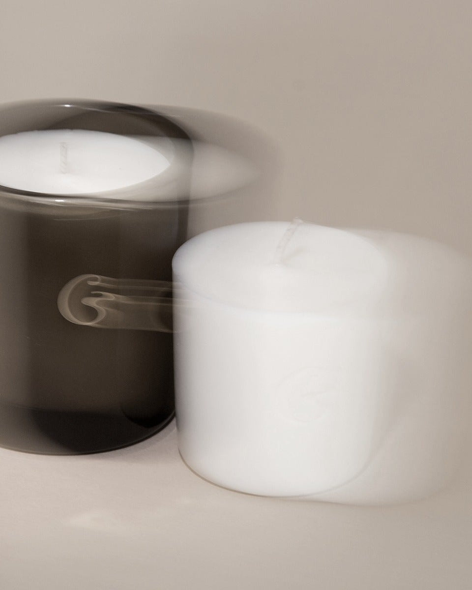 Two candles, one in a glass holder and one plain, on a neutral background