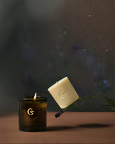 Candle with a logo on a dark background. Next to it is a candle wax refill which is floating on a strange of lavender. 