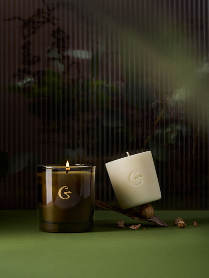 A lit amber candle with a candle refill next to it. set against a dark, atmospheric background with a green surface.
